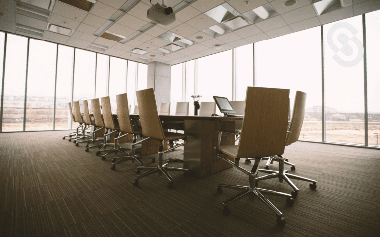 Sala de conferencias vacía con una gran mesa de madera y sillas de oficina giratorias, frente a una pared de ventanas que muestran un paisaje exterior.