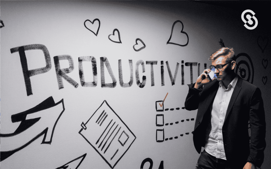 Hombre de negocios hablando por teléfono frente a una pared decorada con la palabra "Productivity" y gráficos relacionados con productividad empresarial y trabajo en equipo.