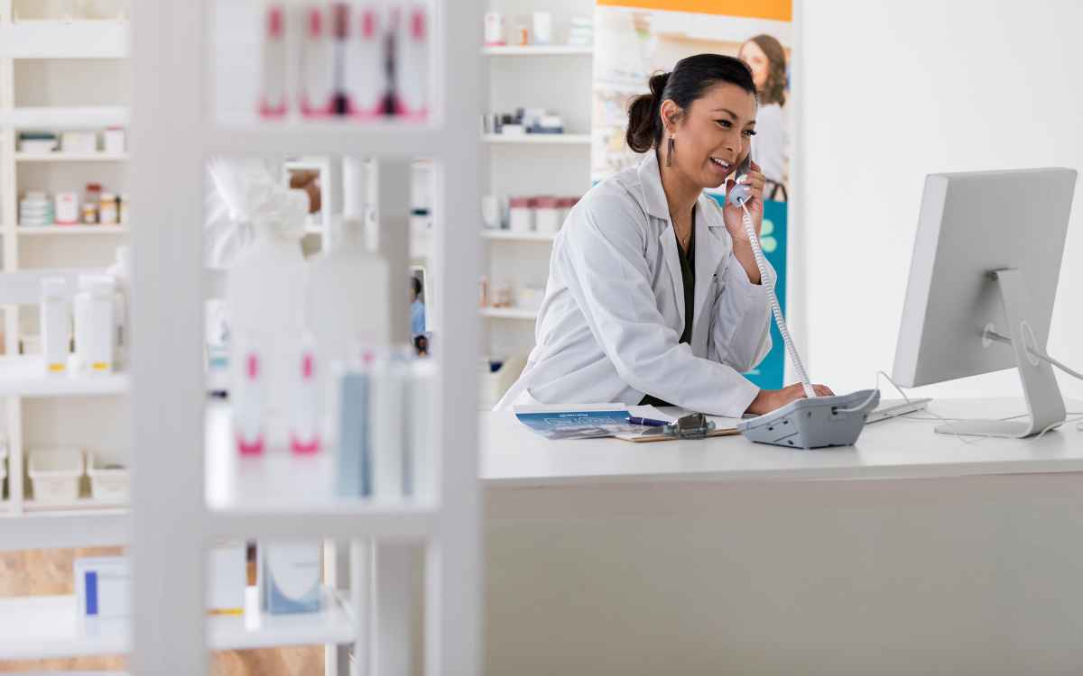 vemos una imagen de una mujer atendiendo el teléfono en una farmacia, mientras observa una computadora, en referencia a los mejores software para farmacias. 