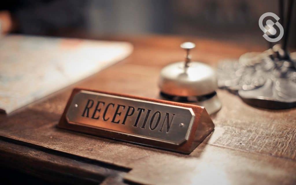 En la imagen se ve la palabra recepción con un pequeño timbre, en una mesa de bienvenida de un hotel. 